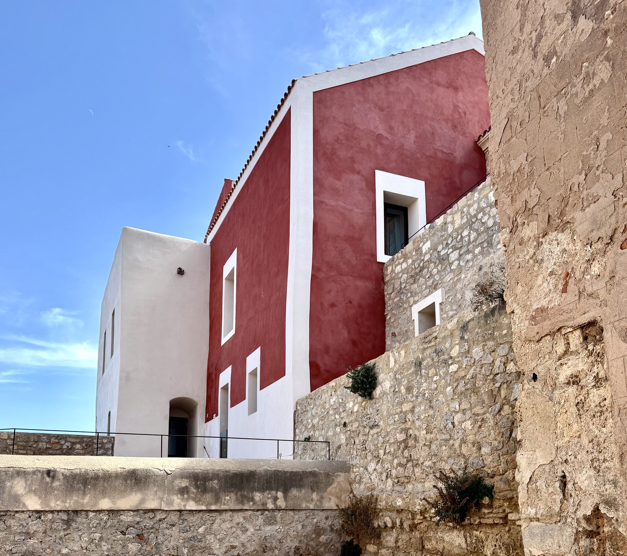 Edificio histórico de color rojo terracota y blanco junto a las murallas de piedra en Dalt Vila, Ibiza.