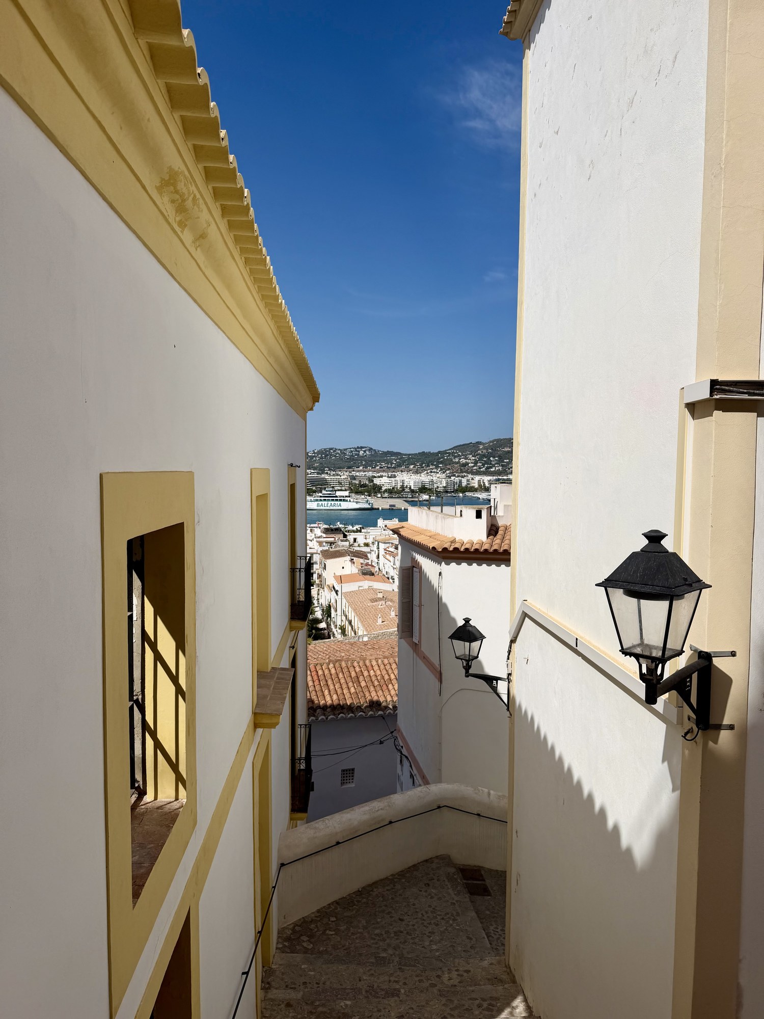 Callejón escalonado empedrado con farolas tradicionales y vistas al puerto de Ibiza y los ferrys.