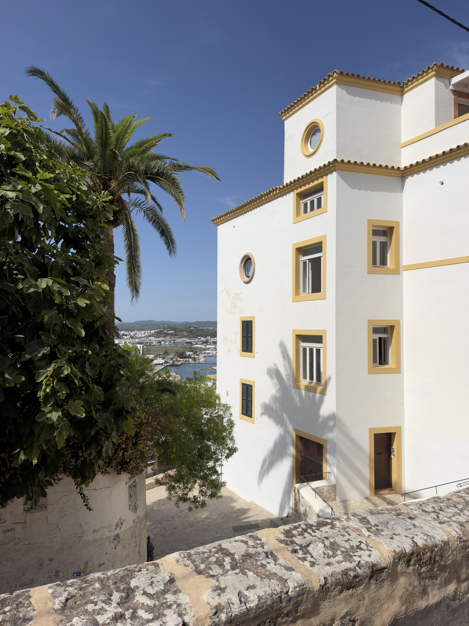 Edificio mediterráneo blanco y amarillo junto a una palmera con vistas panorámicas al puerto de Ibiza.